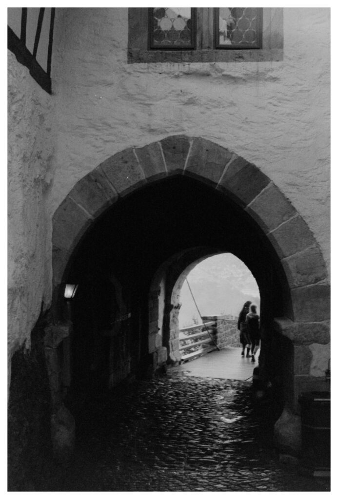 a tunnel in a castle wall, paved with cobblestones reflecting light. outside the tunnel is obscured by fog, two people are just past it
